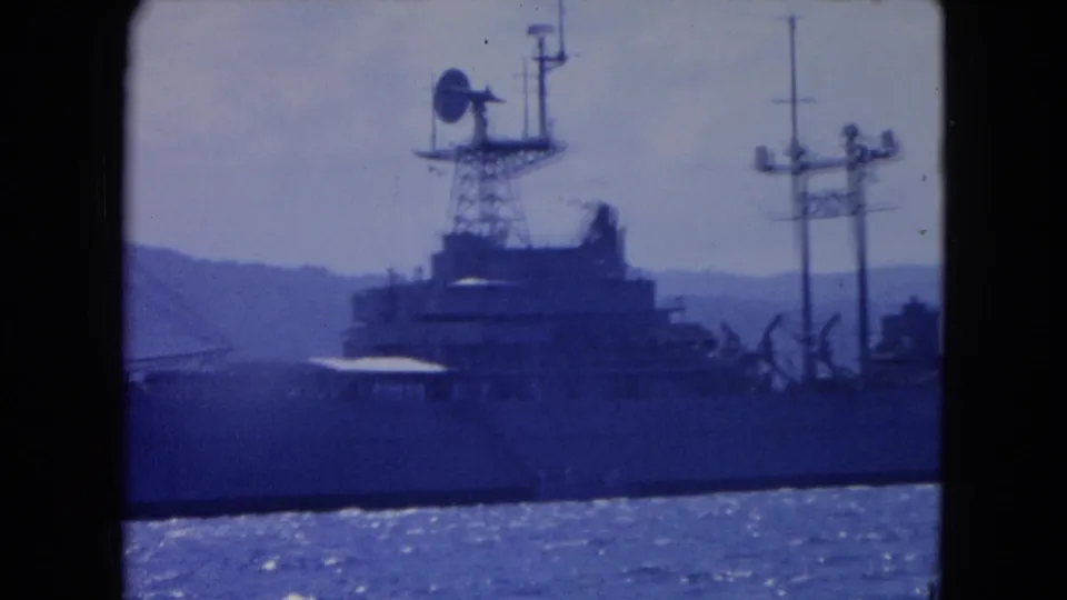 1964 Montego Bay 8mm frame showing the ship superstructure, a dish-shaped apparatus, deck cranes, masts, water, and hills.