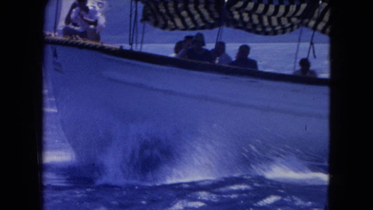 1964 Montego Bay 8mm frame of a white passenger boat cutting through blue water with riders shaded by a striped canopy.