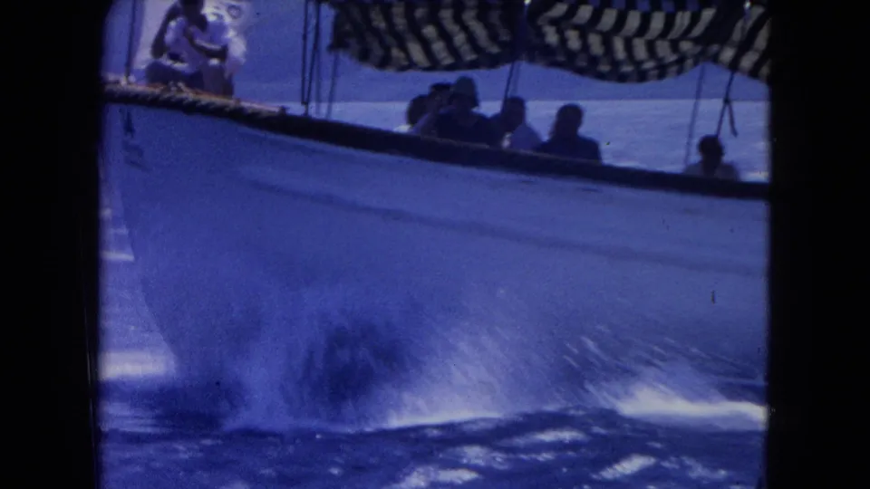 1964 Montego Bay 8mm frame of a white passenger boat cutting through blue water with riders shaded by a striped canopy.