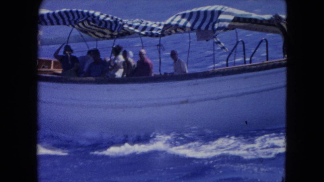 1964 Montego Bay home-movie frame showing a small passenger boat with seated travelers under a striped canopy on blue sea.