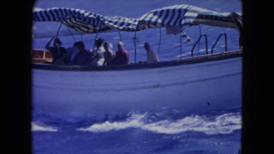 1964 Montego Bay home-movie frame showing a small passenger boat with seated travelers under a striped canopy on blue sea.