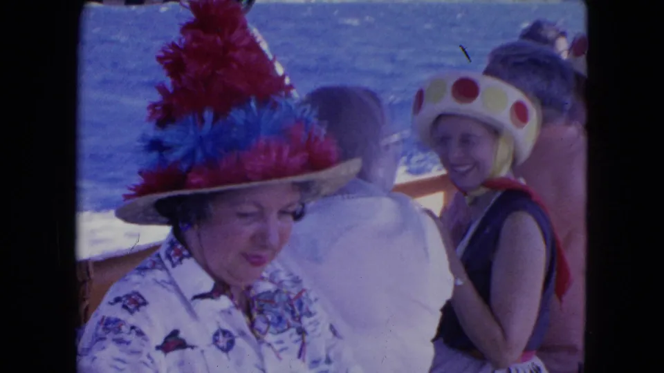 Women on deck in Montego Bay travel footage, including one feathered hat and one polka-dotted headpiece, with water behind them.