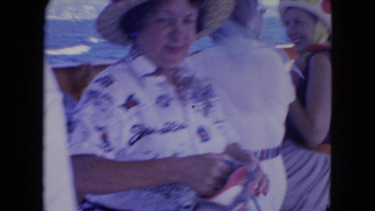 1964 Montego Bay home-movie frame showing adults on a boat deck, a feathered hat, a Jamaica shirt, and blue sea in the background.