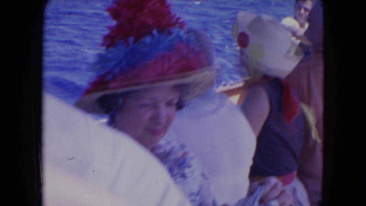 1964 Montego Bay frame with a smiling woman in a colorful feathered hat among adults on a sunlit boat deck beside the sea.