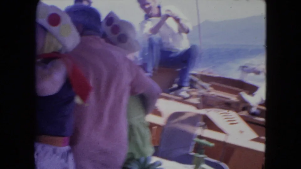 1964 Montego Bay frame showing adults in novelty hats on deck, a man near the rail, sea, and distant shoreline.