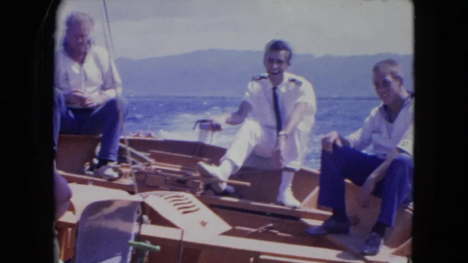 The uniformed man smiles while seated on the wooden boat in Montego Bay, with passengers and choppy water visible.