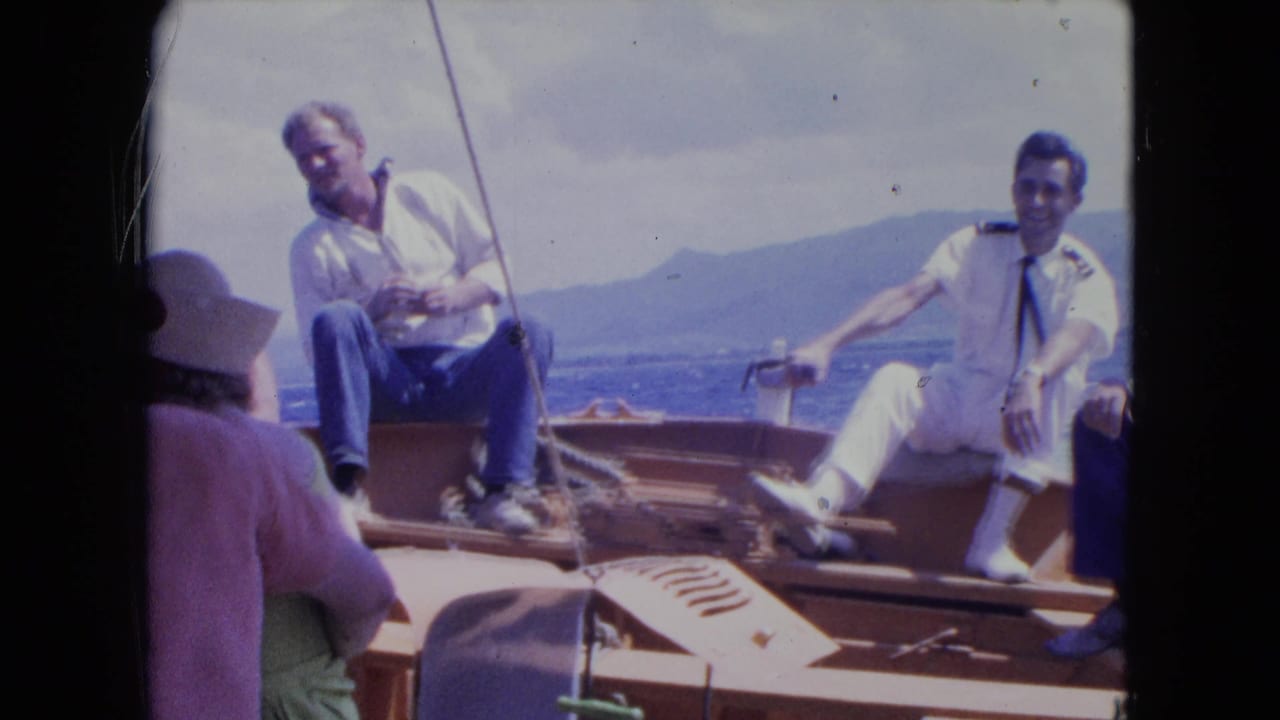 1964 Montego Bay home-movie frame showing adult men on a wooden boat, including one in a white uniform, with coastline behind.