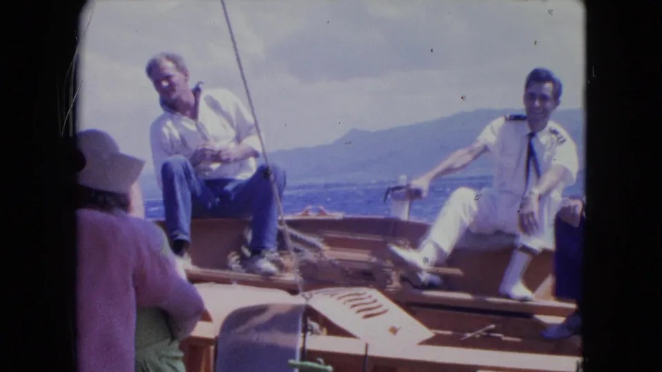 1964 Montego Bay home-movie frame showing adult men on a wooden boat, including one in a white uniform, with coastline behind.