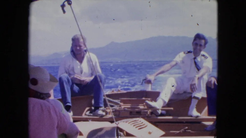 Montego Bay boat scene with two seated adult men, one in a white uniform, on deck against blue water and distant hills.