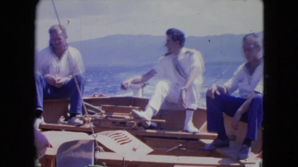 1964 Jamaica home-movie frame showing three adult men seated on a small wooden boat, with one uniformed man turned sideways.