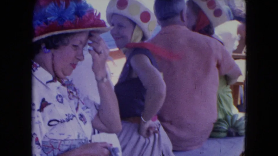 1964 Montego Bay home-movie frame with women in elaborate vacation hats beside a shirtless man on a crowded boat deck.