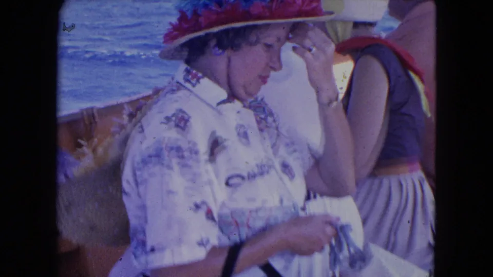 1964 Montego Bay frame showing a woman in a large flowered hat and patterned shirt beside another vacationer on a wooden boat.