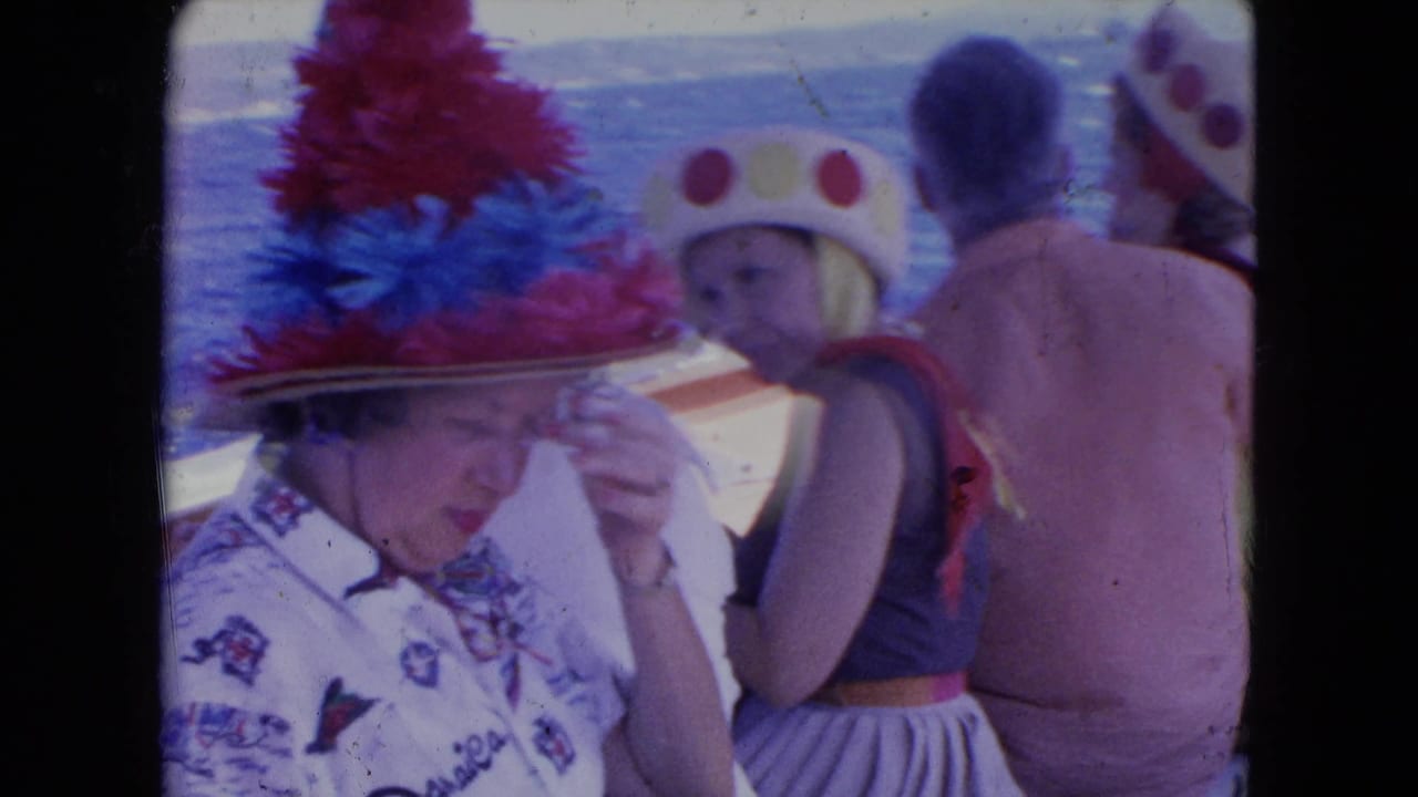 Passengers sit on a boat as a woman in a bright feathered hat adjusts her brim.