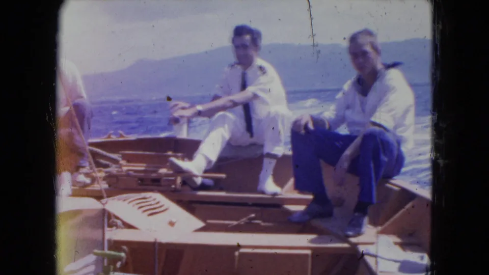 Two men sit casually on a wooden boat deck in supported Montego Bay footage, with bright water and distant hills behind them.