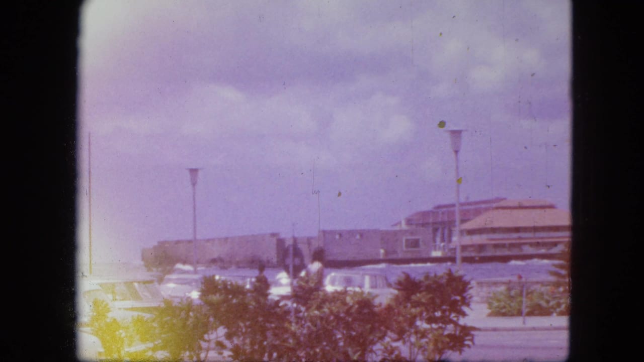 1964 Nassau waterfront view with a tan-roofed seaside building, low shrubs, parked cars, lamps, and aged 8mm color cast.