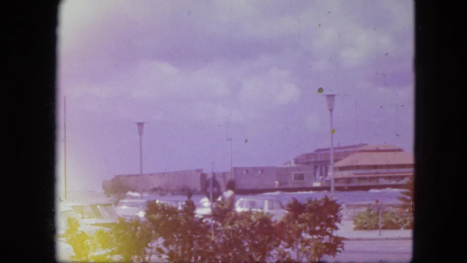 1964 Nassau waterfront view with a tan-roofed seaside building, low shrubs, parked cars, lamps, and aged 8mm color cast.