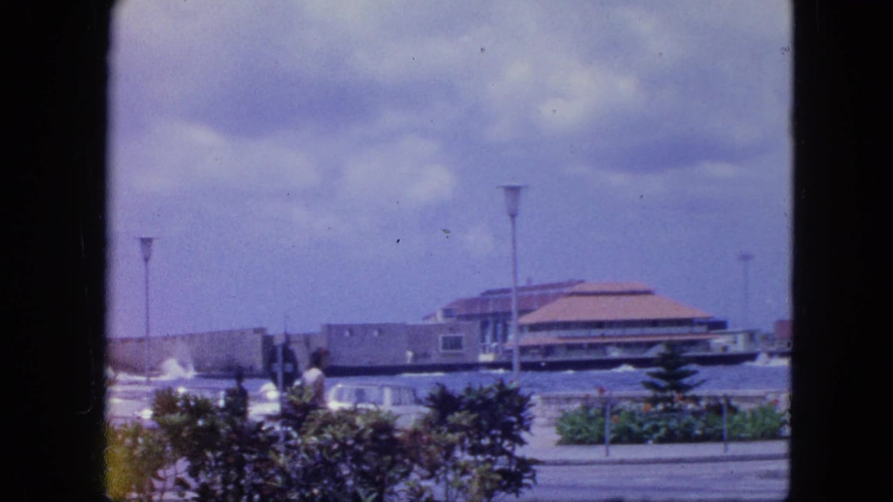Nassau seaside building in 1964 seen across a promenade, with pedestrians, parked cars, shrubs, lamps, water, and cloudy sky.