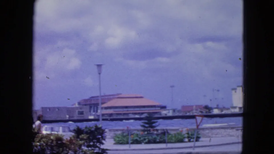 1964 Nassau waterfront promenade with a tan-roofed building, low plantings, a lamppost, cloudy sky, and a partly visible pedestrian.