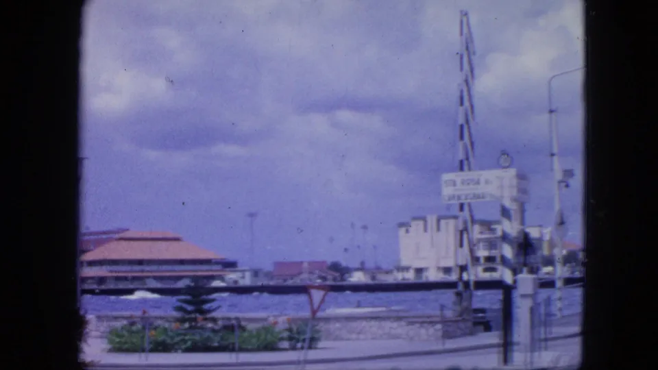 Nassau waterfront view in 1964 with the tan-roofed building, a visible sign at right, lampposts, shrubs, road edge, and cloudy sky.