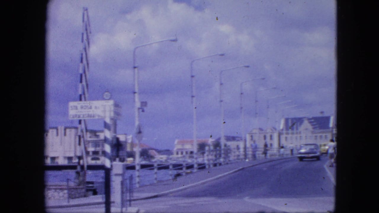 Roadway across a bridge with a classic car, harbor railings, sign, and waterfront buildings.