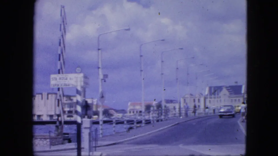Nassau bridge roadway in 1964 with a classic car approaching, harbor railings, a visible sign, and red-roofed buildings in the distance.