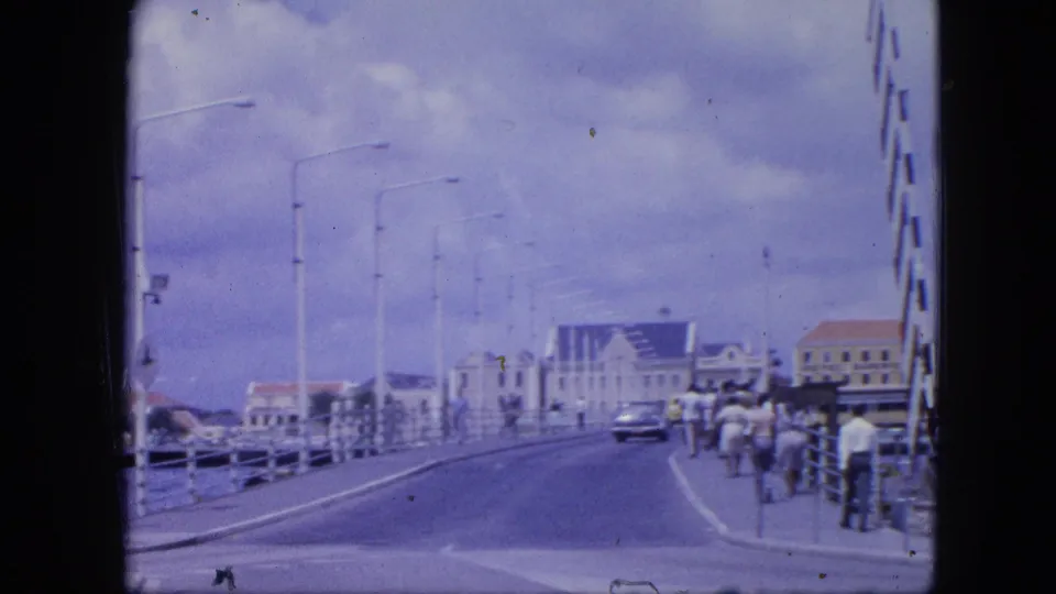 1964 Nassau waterfront bridge scene showing pedestrians walking along the right side, a classic car ahead, street lamps, and harbor buildings.