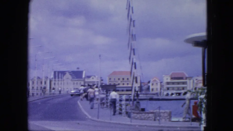 Nassau bridge and waterfront in 1964 with pedestrians at the roadside, a classic car ahead, water to the right, and low buildings beyond.