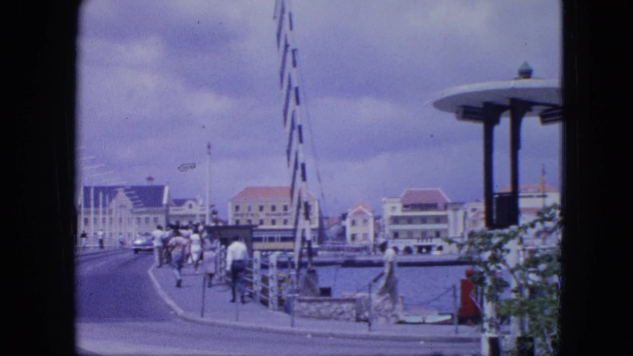 1964 Nassau waterfront view with people on the promenade, harbor railings, red-roofed buildings, and a modernist circular-roof pavilion.