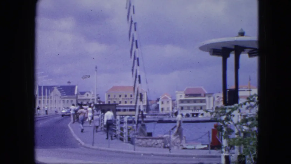 1964 Nassau waterfront view with people on the promenade, harbor railings, red-roofed buildings, and a modernist circular-roof pavilion.