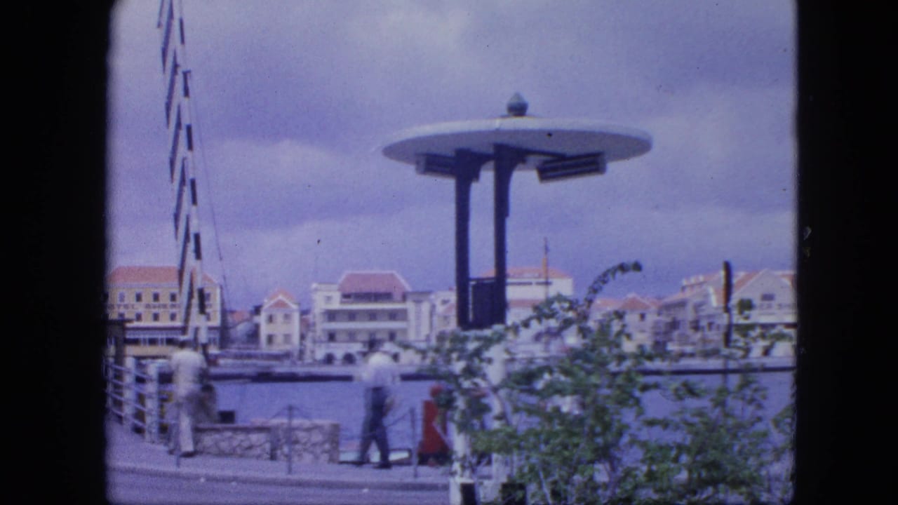 People walk near harbor railings as waterfront buildings and a circular-roof pavilion appear across the frame.