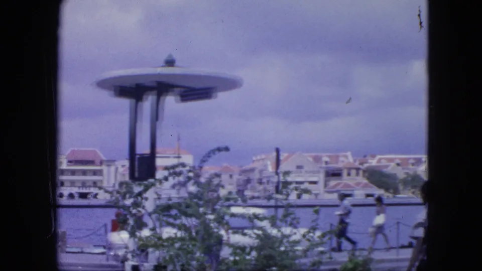 1964 Nassau harborfront scene with a modernist circular-roof pavilion, pedestrians on the promenade, water, and red-roofed buildings.