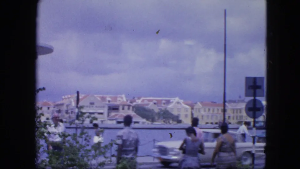 1964 Nassau waterfront road with a passing car, pedestrians, harbor railings, traffic signs, and red-roofed buildings in the background.