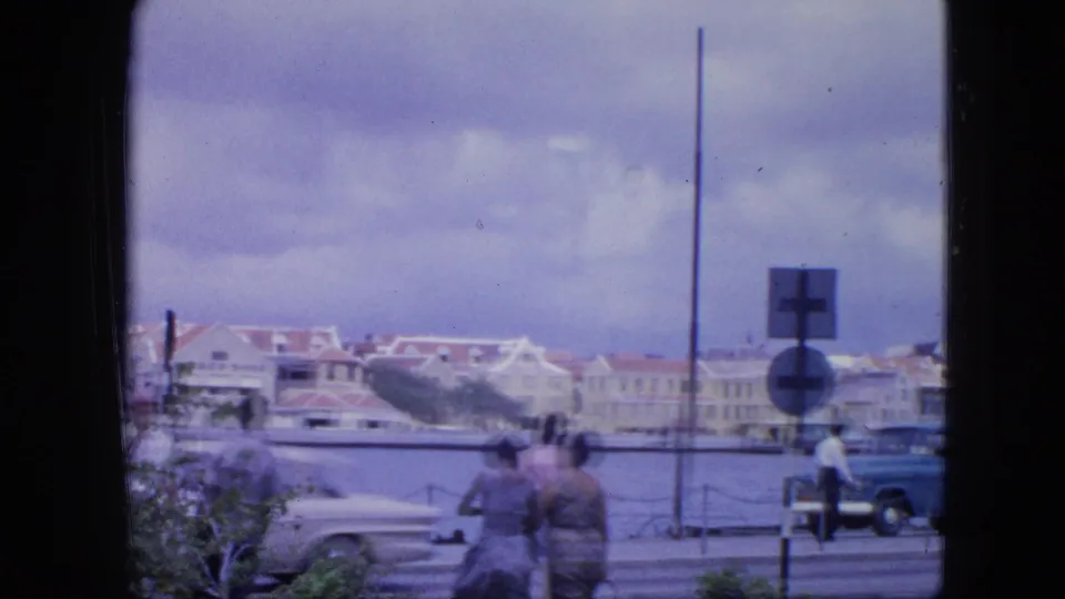 1964 Nassau waterfront street view with classic cars, pedestrians, harbor water, and pale buildings under a cloudy sky.