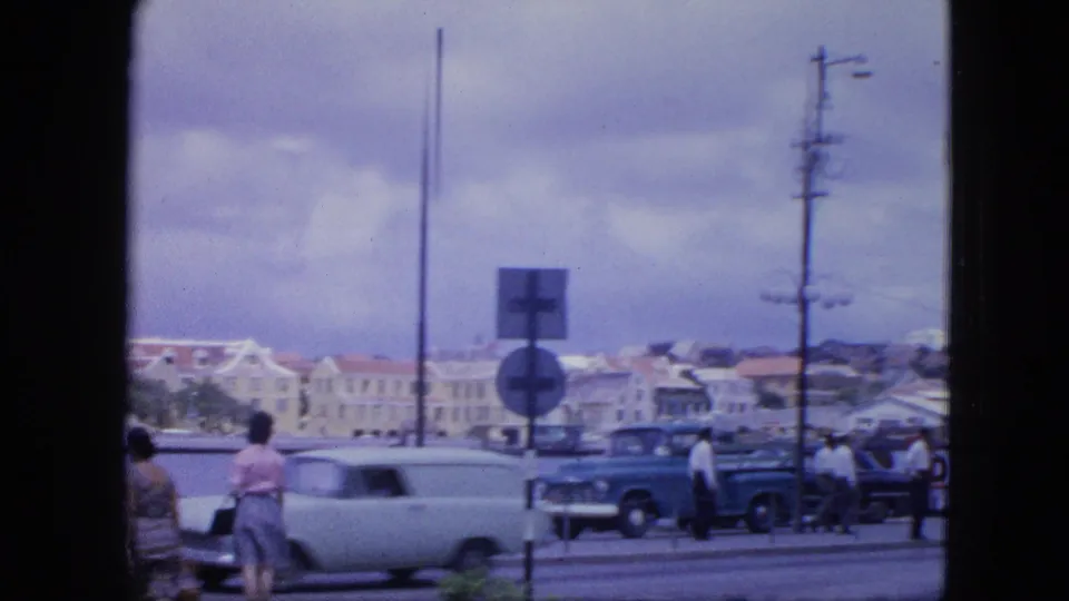 Nassau waterfront traffic in 1964, with a light-colored classic car, pedestrians, road signs, and harbor buildings in the background.