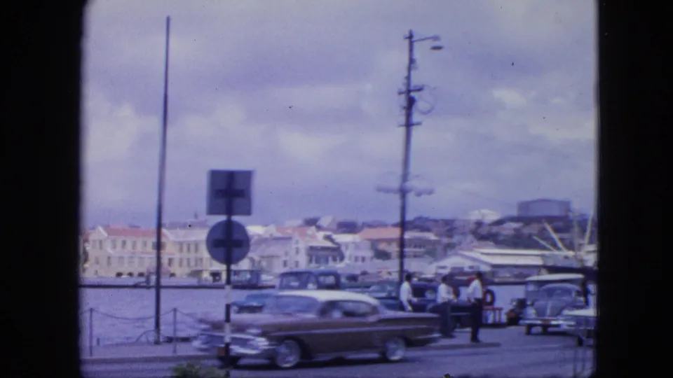 1964 Nassau harbor road scene with a darker classic car, standing pedestrians, road signs, water, and waterfront buildings.