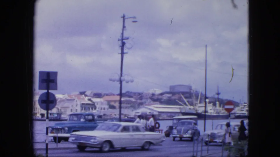 White classic car on a 1964 Nassau waterfront street, with pedestrians, parked vehicles, harbor water, signs, and utility poles visible.