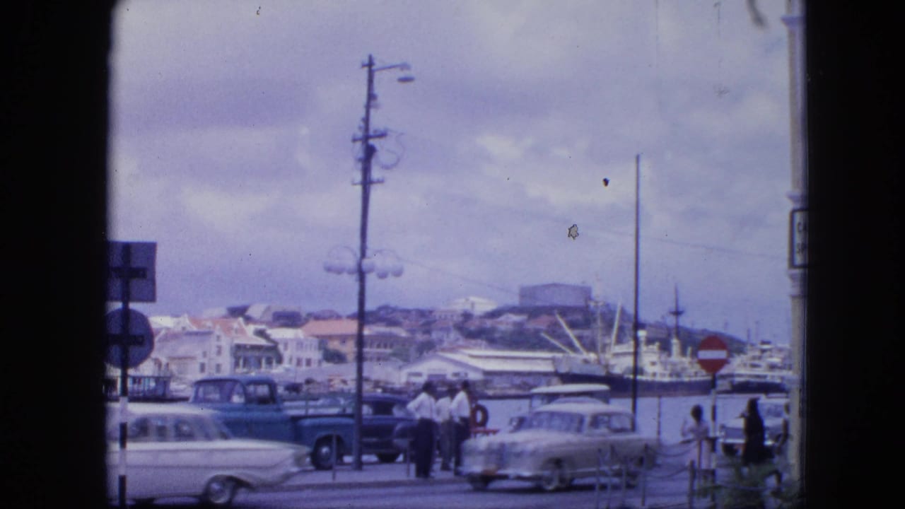1964 Nassau waterfront street scene with pedestrians, mid-century cars, docked ships, hillside buildings, and traffic signs.