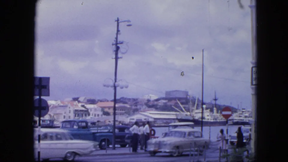 1964 Nassau waterfront street scene with pedestrians, mid-century cars, docked ships, hillside buildings, and traffic signs.