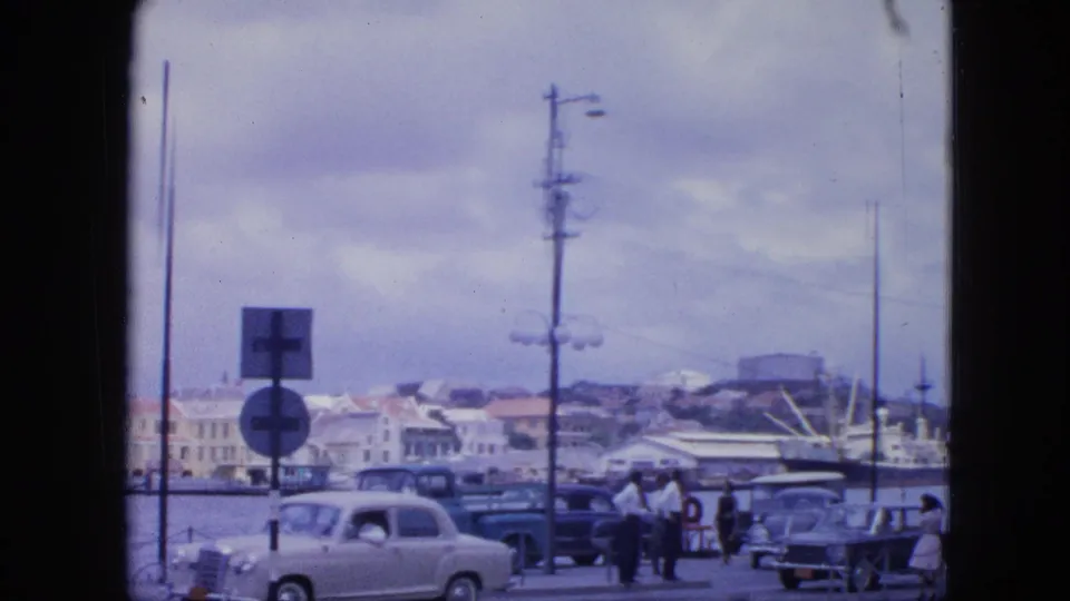 Nassau harbor street in 1964 with a pale sedan, pedestrians, parked cars, docked vessels, signs, and waterfront buildings.