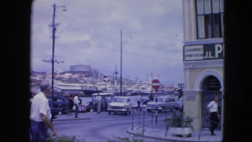 1964 Nassau sidewalk view showing pedestrians, mid-century cars, harbor ships, road signs, and a partial storefront sign.