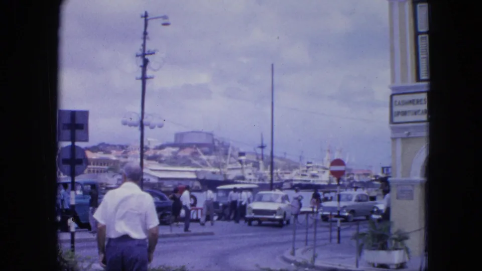 Nassau harbor street scene from 1964 with a man seen from behind, pedestrians, cars, ships, signs, and a storefront edge.