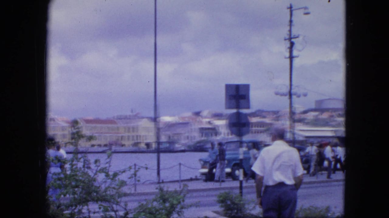 Waterfront street seen through a dark frame, with a man, pedestrians, parked cars, harbor water, and buildings.