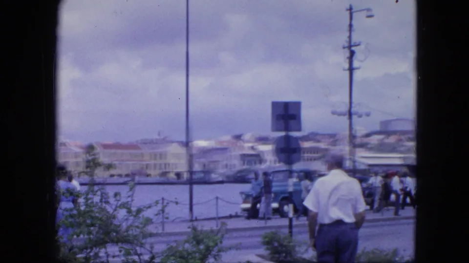 Nassau harbor street in 1964 seen through a dark window frame, showing a man in white, pedestrians, cars, signs, water, and low buildings.