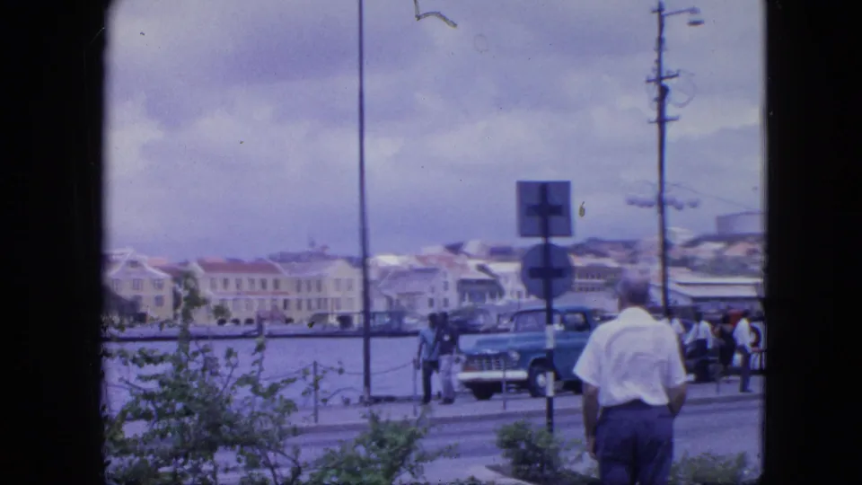 1964 Nassau waterfront street with pedestrians, period cars, road signs, a utility pole, harbor water, and pastel buildings under a cloudy sky.