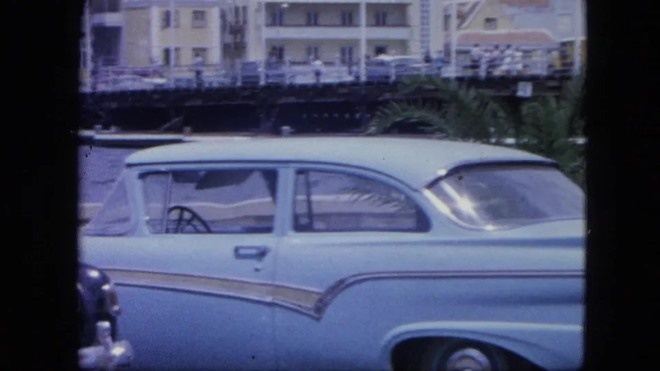 Close view of a light blue period car on the Nassau waterfront in 1964, with railings, harbor buildings, parked cars, and palm foliage behind it.