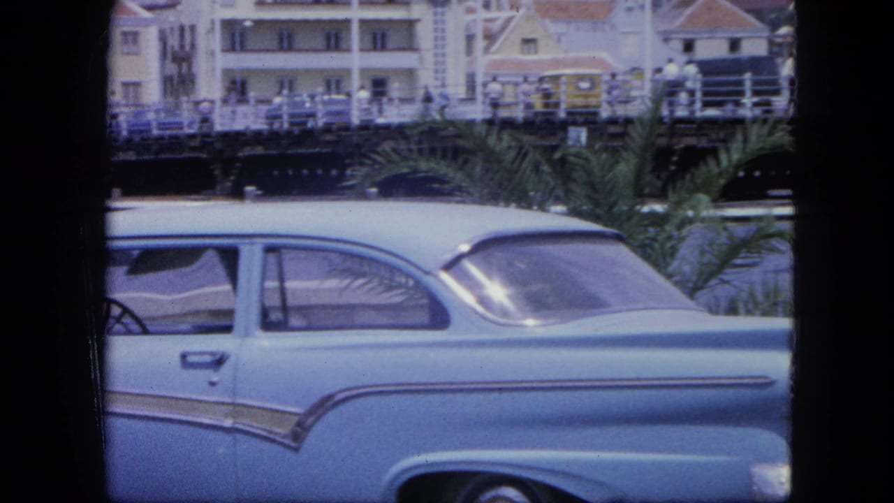 1964 Nassau waterfront view with a light blue sedan, palm fronds, water, bridge railings, and colonial-style buildings behind it.