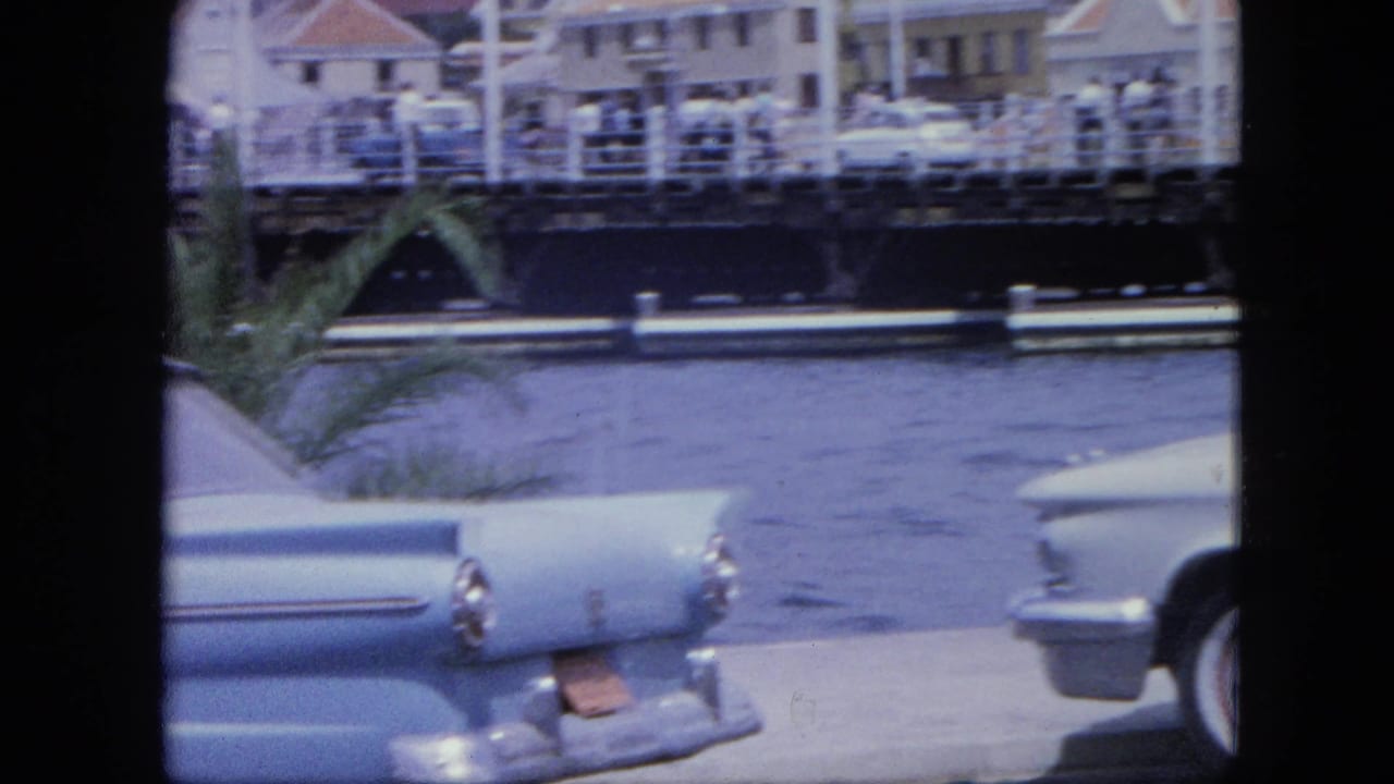 Parked car beside water with bridge railings, buildings, and pedestrians in the background.