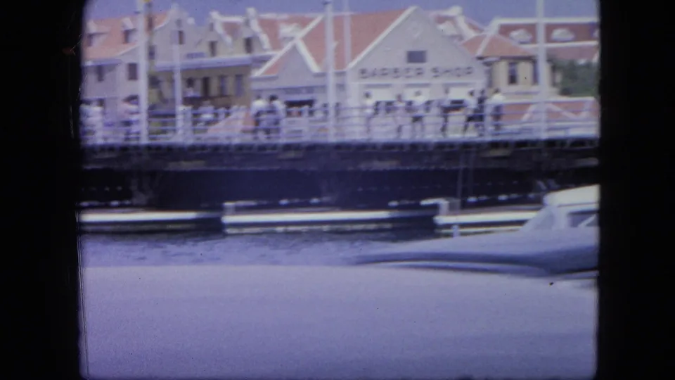 1964 Nassau waterfront view with car roofline in front, water and bridge railings beyond, and pedestrians near colonial-style buildings.