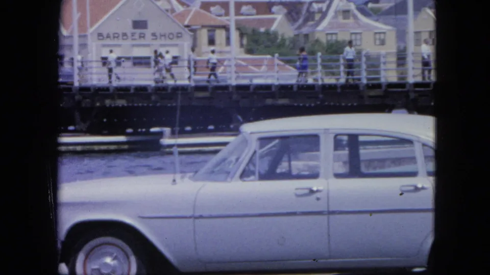 Nassau waterfront frame with a white vintage sedan, bridge railings, pedestrians, and a visible barber shop sign on a building.