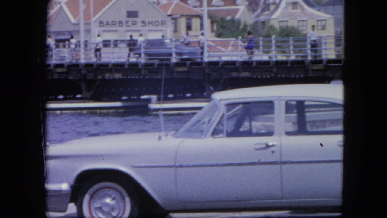 White sedan beside water with pedestrians on a bridge and a barber shop sign across the waterfront.